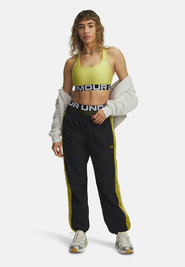 Geel sportbeha met zwarte band, zwarte joggingsbroek met gele zijstrepen, witte jas over de schouders gedrapeerd, grijze sneakers met accenten.