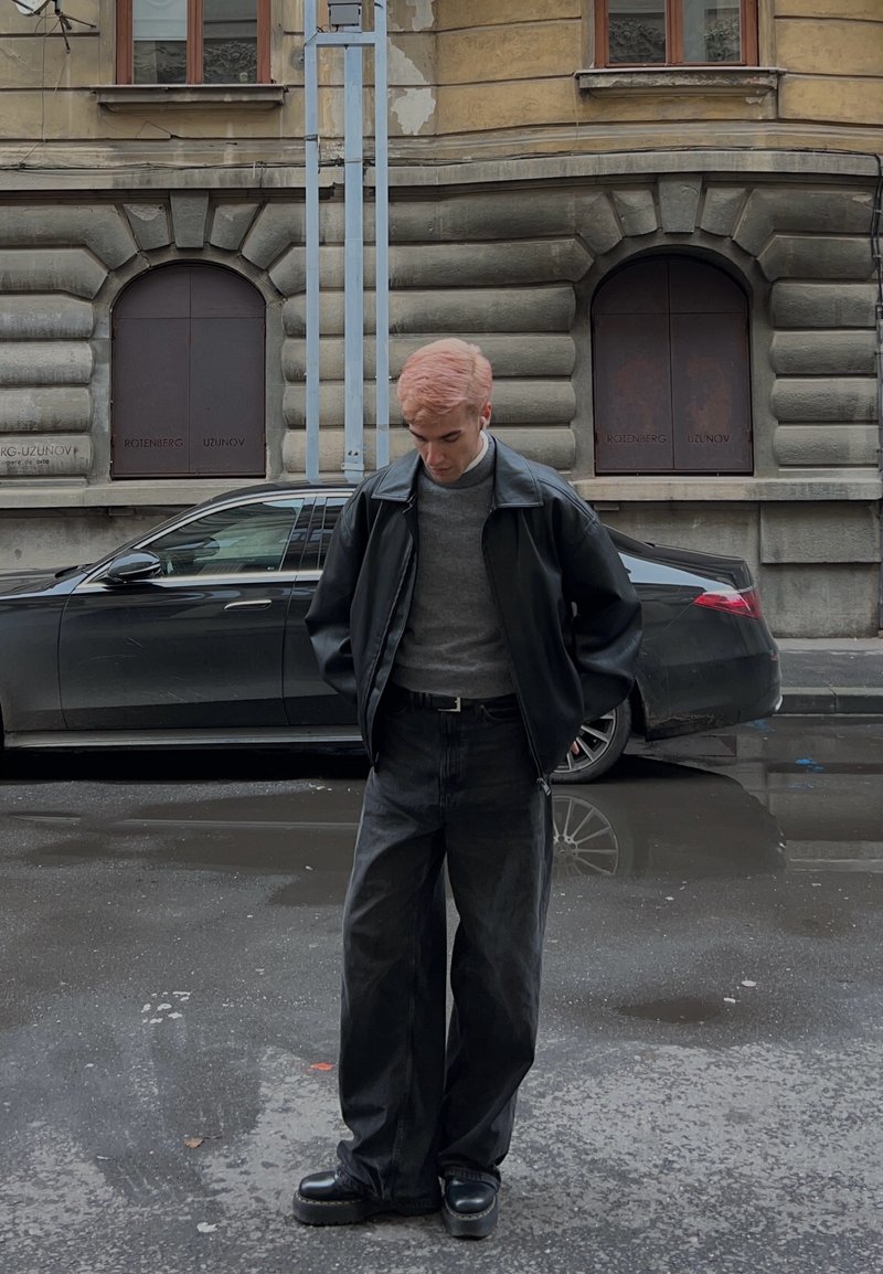 Giacca di pelle nera sopra un maglione grigio, abbinata a jeans neri larghi e scarpe platform nere. Capelli rosa chiaro e sfondo neutro.