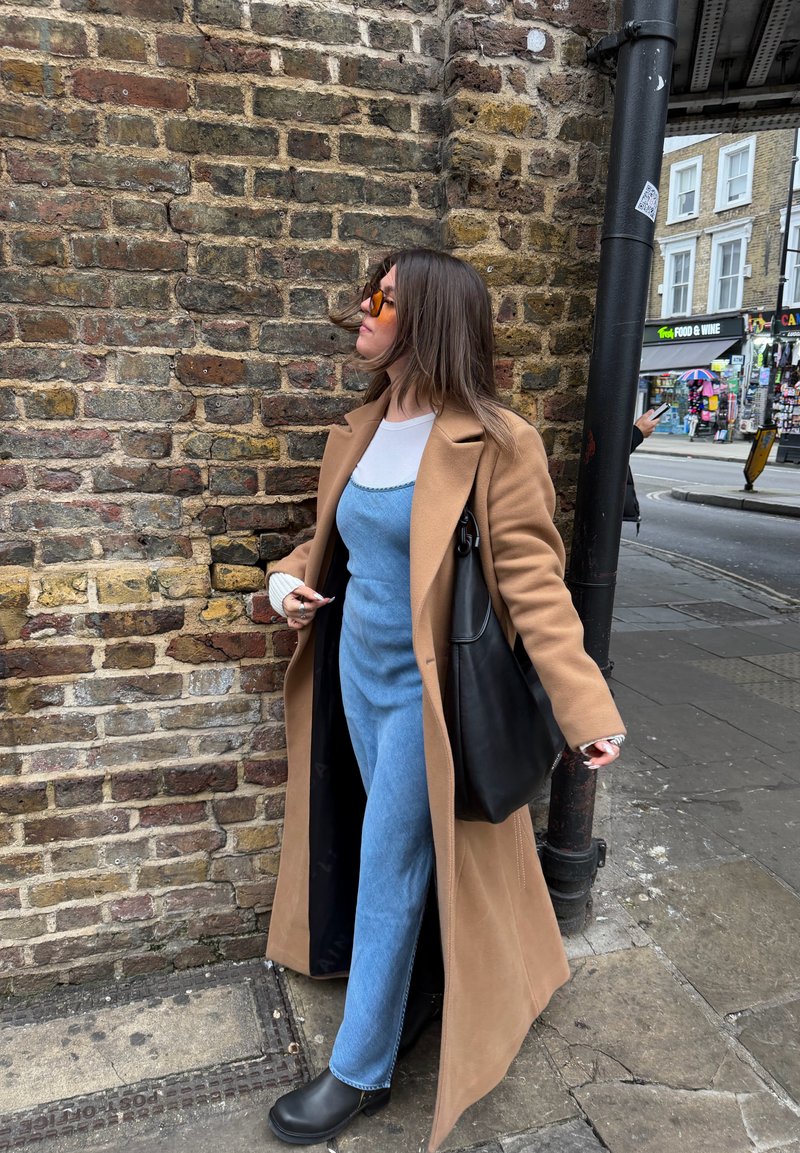 Γυναίκα που φοράει μακριά καμηλό παλτό και μπλε φόρεμα, στέκεται δίπλα σε τοίχο από τούβλα σε μια αστική οδό, κρατώντας μια μεγάλη μαύρη τσάντα.