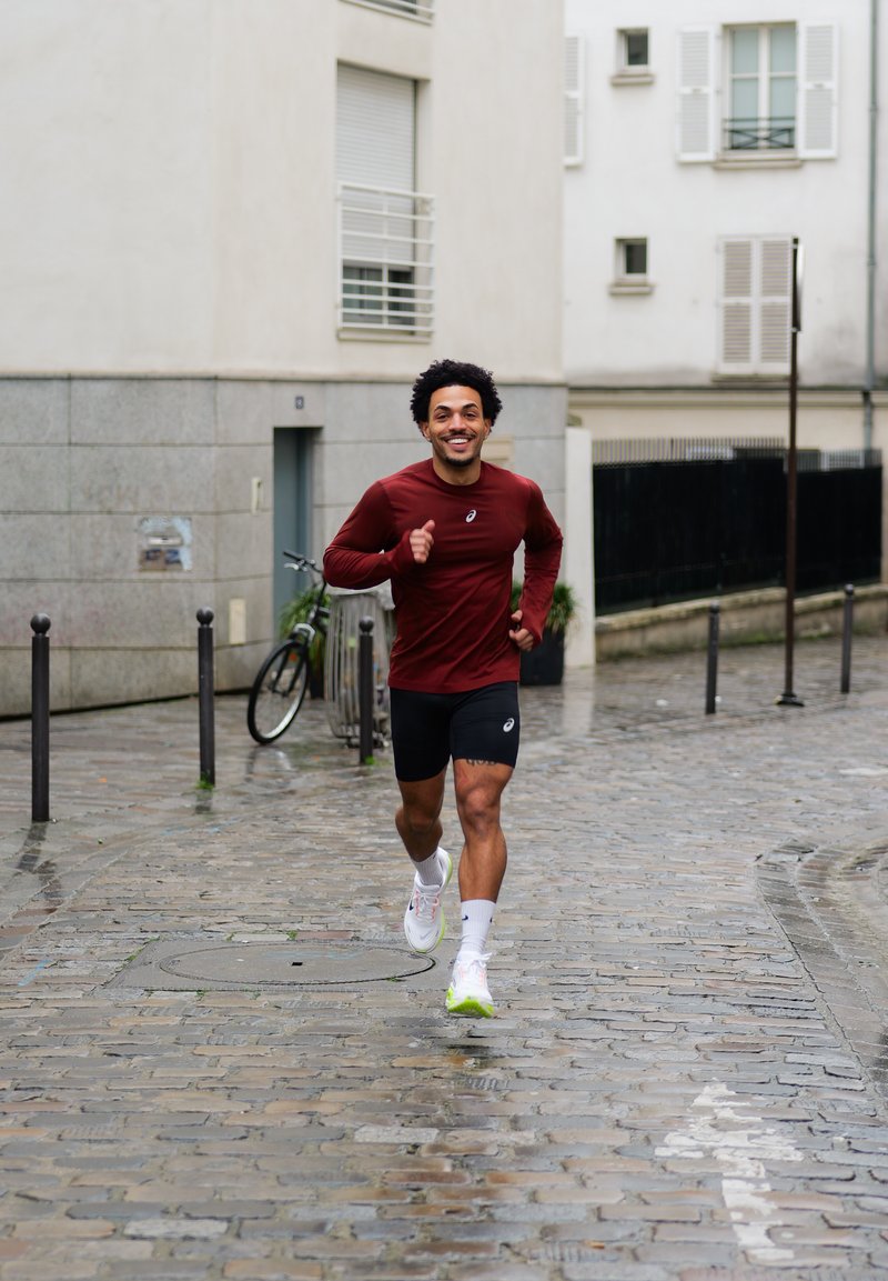 Άνδρας που φοράει μακρυμάνικο πουκάμισο μπορντό και μαύρα σορτς τρέχει σε υγρό πλακόστρωτο δρόμο σε αστική περιοχή με ποδήλατο στο παρασκήνιο.