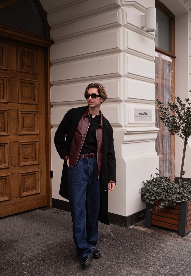 Abrigo negro sobre una chaqueta de cuero burdeos, polo negro y jeans altos de talle azul. Zapatos negros completan el atuendo. Fondo de puerta de madera y vegetación.