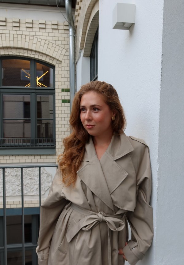 Vaaleanbeige trench-mantel, jossa on vyöllinen vyötärö ja leveä kaulus. Sujuvantuntuinen kangas, pitkät hihat ja sivutaskut. Taustalla on suuret ikkunat.