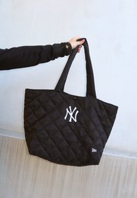 Mão segurando uma bolsa tote acolchoada preta com logo branco de NY e alças almofadadas em frente a uma parede de fundo texturizada clara.