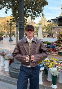 Giacca corta in pelle marrone con cuciture a contrasto, chiusura con bottoni e colletto alto. Abbinata a un maglione verde con fantasia e jeans larghi.