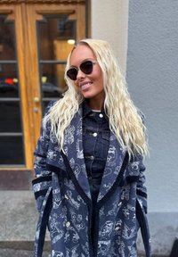 Μπλε παγιέτα με περίπλοκα σχέδια, φορεμένο πάνω από τζιν ρούχα. Μακριά, κυματιστά ξανθά μαλλιά, σκούρα γυαλιά ηλίου και ουδέτερο φόντο.