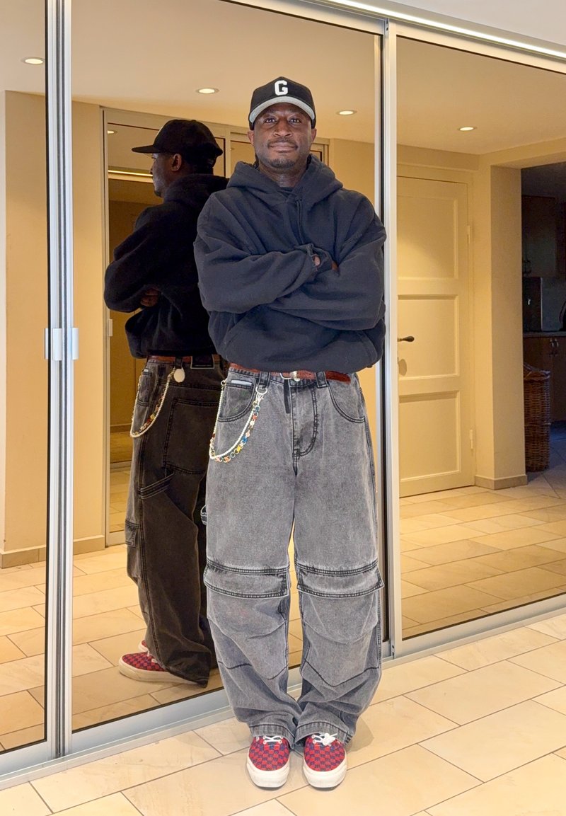 Homme portant un sweat à capuche noir, un jean baggy gris avec chaîne, des baskets rouges à carreaux et une casquette noire, debout les bras croisés devant des portes de placard miroir.