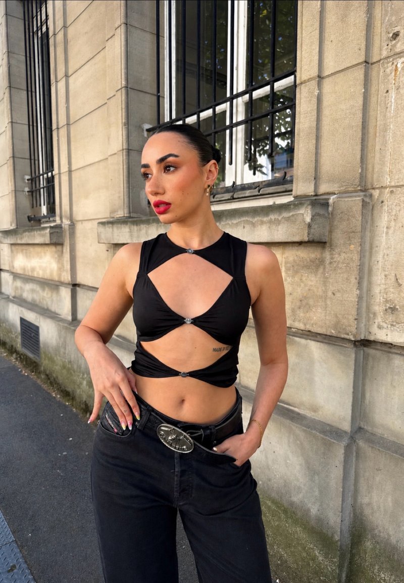 Femme aux cheveux tirés en arrière porte un haut noir avec découpes et un jean noir avec une grande boucle de ceinture, debout contre un bâtiment en pierre avec des fenêtres grillagées.