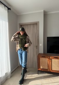 Femme portant un pull à motifs, une écharpe verte, une casquette de baseball, un jean et de hautes bottes vertes, debout à l'intérieur près d'un meuble TV en bois et d'une porte fermée.