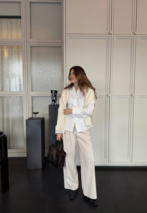 Woman in white blouse and cream pants holding a woven black bag, standing indoors by textured glass panels and tall grey pedestals.