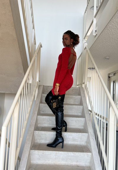 Vestido rojo de manga larga con la espalda al aire, combinado con botas de cuero negras hasta la rodilla. Sostiene un pequeño bolso negro con acentos dorados.