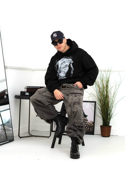 Sudadera negra con estampado gráfico, pantalones cargo grises con múltiples bolsillos y botas negras con cordones. Entorno interior minimalista.