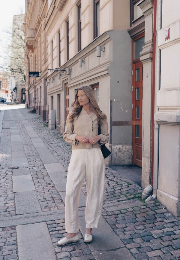 Nuori nainen beige takki ja valkoiset housut päällä seisoo mukulakivikadulla lähellä historiallisia rakennuksia, katsoo sivulle kämmenet yhteen kiedottuna.