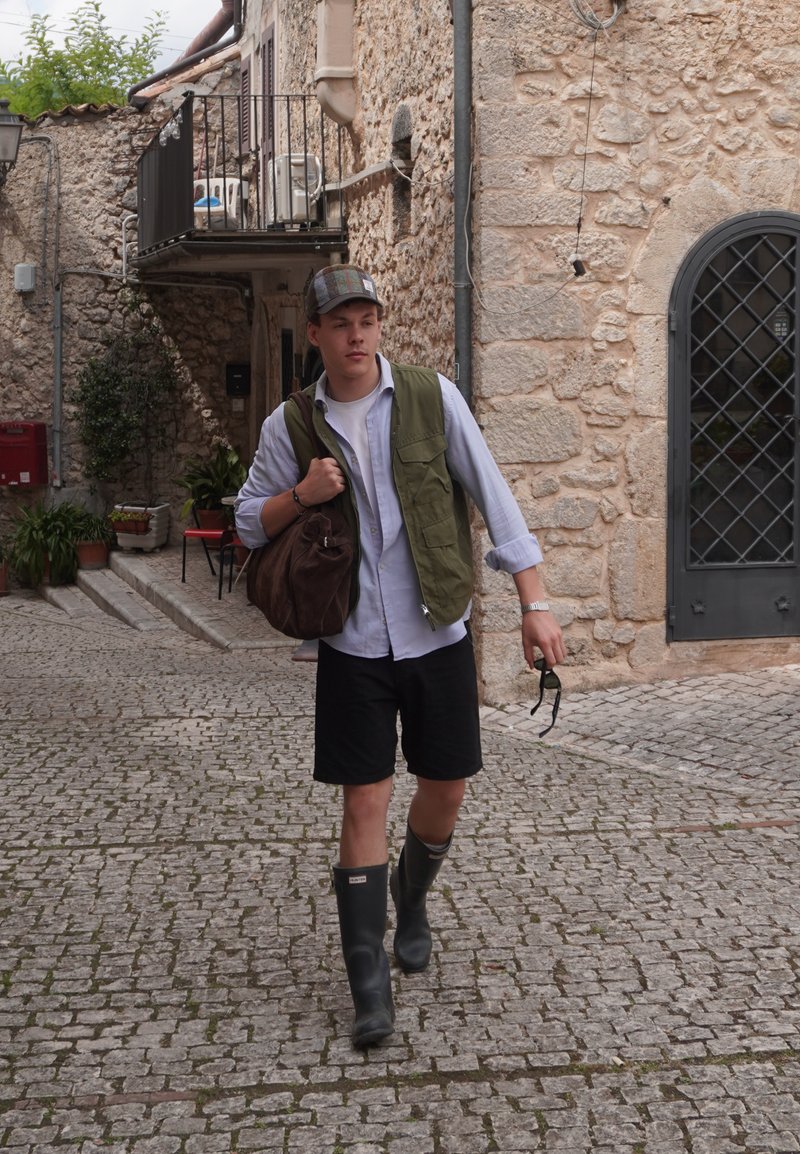 Jeune homme portant une casquette, un gilet vert, un short noir et des bottes en caoutchouc marche sur une rue pavée en tenant des lunettes de soleil et un sac marron.