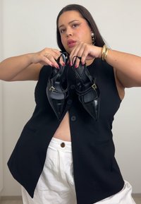 Melni lakādas spicpapēžu kurpes ar dubultajām siksnām un zelta detaļām, ko tur persona, kura valkā melnu veste un baltas bikses.