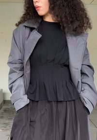 Abrigo exterior gris con cuello, superpuesto a una blusa peplum negra con pliegues, combinado con una falda negra brillante. Texturas suaves y diseño ajustado.
