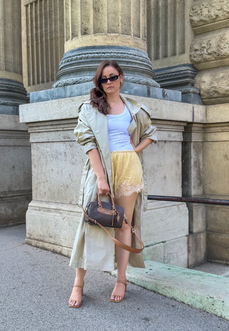 Femme portant des lunettes de soleil, un trench léger, un débardeur blanc, un short jaune bordé de dentelle, tenant un sac à main gris, debout près de colonnes en pierre.