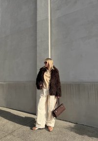 Rjav umetni krzneni plašč, smetanast pulover, široke smetanaste hlače, rjava torbica in bež čevlji. Stoji ob teksturirani sivi steni.
