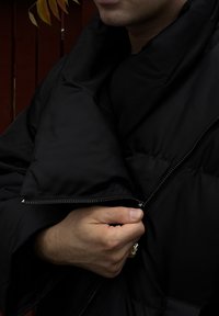 Črna pufasta jakna z visokim ovratnikom, ki ima sprednji zadrgo in mehak, teksturiran material. Model drži zadrgo z eno roko.