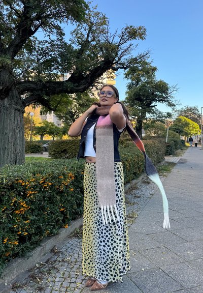 Bufanda larga multicolor con un degradado de oscuro a claro, llevada sobre una camiseta blanca y una falda amarilla con lunares. Ambiente al aire libre.