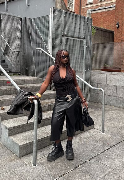 Top negro sin mangas, culottes de cuero de cintura alta, botas negras gruesas, hebilla de cinturón de plata. Escaleras de piedra con barandilla de metal y fondo texturizado.