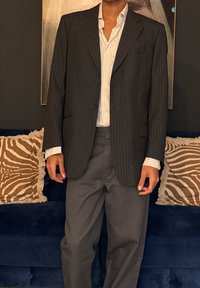 Άνδρας που φοράει μαύρο σακάκι με ρίγες πάνω από λευκό πουκάμισο με ρίγες και σκούρο παντελόνι, στέκεται μπροστά από έναν μπλε καναπέ με μαξιλάρια με ζέβρα σχέδιο.