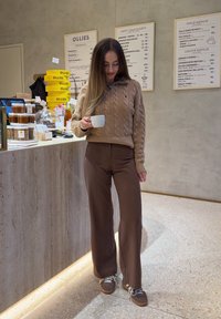 Žena s dlouhými vlasy, oblečená v hnědém svetr s copánkovým vzorem a hnědých kalhotách, držící bílý hrnek, stojící uvnitř kavárny blízko pultu.