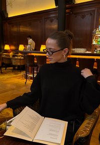 Pull noir en maille à col haut, lisant un menu à une table, entouré d'un éclairage chaleureux et de mobilier en bois dans un cadre élégant.