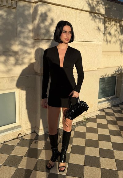 Mujer con cabello corto y negro, vestida con un mini vestido negro de manga larga y botas hasta la rodilla con punta abierta, sosteniendo un pequeño bolso negro al aire libre.