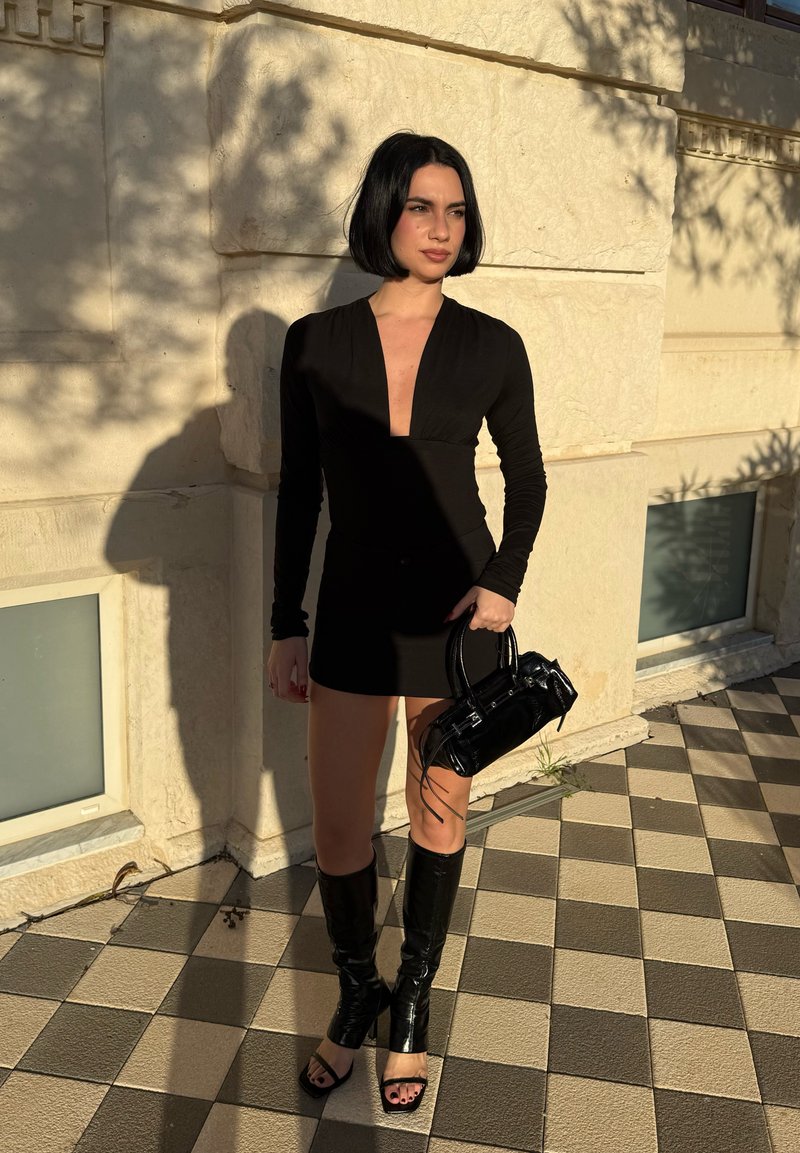 Femme aux cheveux courts noirs portant une mini robe noire à manches longues et des bottes ouvertes aux genoux, tenant un petit sac à main noir en plein air.