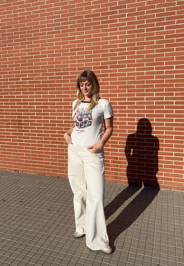 Mujer con pantalones blancos y camiseta gráfica está con las manos en los bolsillos contra una pared de ladrillos rojos, proyectando una sombra en la pared detrás de ella.
