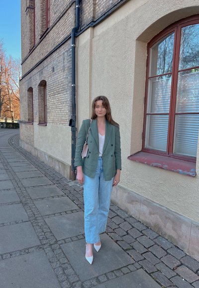 Blazer verde de doble pecho, blusa blanca, jeans rectos de mezclilla azul claro, tacones blancos de punta, bolso negro con cadena, ambientación en la acera.