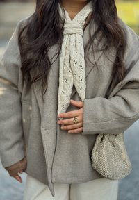 Bežový nadmerný vlnený kabát, krémový šál s textúrou a vzorovaná hnedá taška. Doplnky zahŕňajú zlatý prsteň s dekoratívnym vzorom.