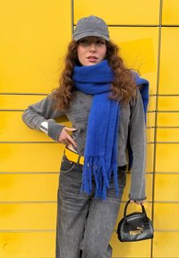 Pulôver cinza, cachecol azul com franjas, mala preta com logotipo dourado, cinto amarelo, jeans cinza e um boné cinza contra um fundo amarelo brilhante.