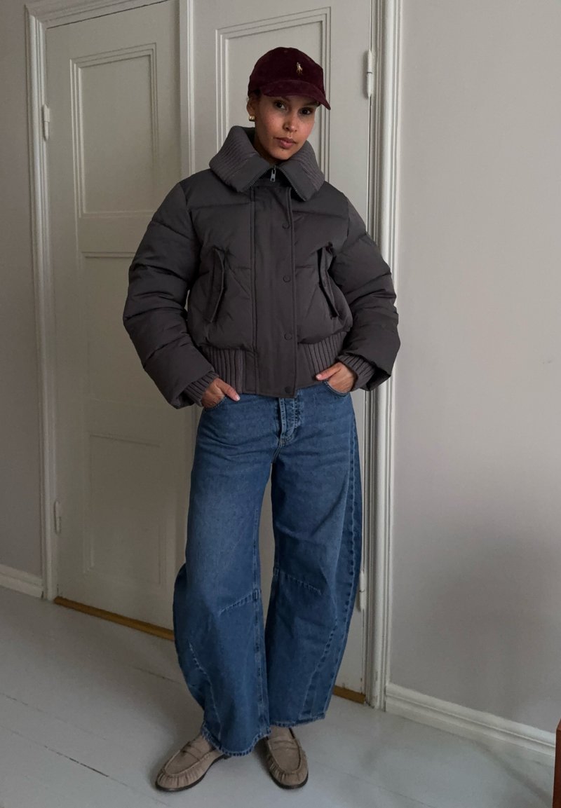 Femme portant une doudoune sombre, un jean large bleu, des mocassins beige et une casquette de baseball bordeaux, debout à l'intérieur près de portes blanches.