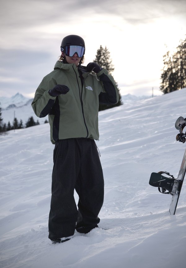 Osoba u zelenoj jakni, crnoj kacigi i naočalama stoji na snježnom terenu pored snowboarda zabodenog u snijeg.