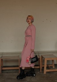 Vestido rosa de manga larga con relieve y cinturón. Botas de plataforma negras con suelas gruesas. Bolso negro con charms. Fondo neutro y muebles de madera.