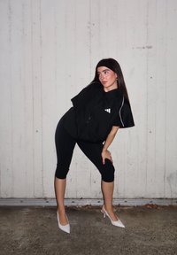 Fekete, túlméretezett, rövid ujjú cipzáras ing fehér logóval és díszítő csíkkal; fekete, felhajtott leggingssel és fehér magassarkúval párosítva.