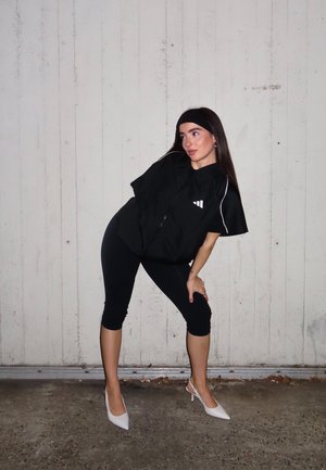 Camisa negra extragrande con cierre y mangas cortas, con un logotipo blanco y ribetes; combinada con leggings negros recortados y tacones blancos.