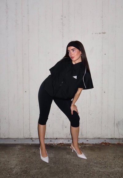 Camisa negra extragrande con cierre y mangas cortas, con un logotipo blanco y ribetes; combinada con leggings negros recortados y tacones blancos.