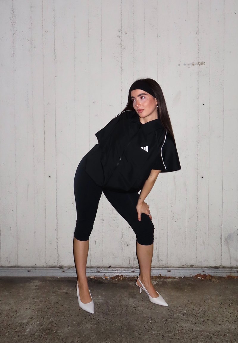 Camisa preta oversized com fecho de correr e mangas curtas, com um logótipo branco e ribetes; combinada com leggings pretas curtas e saltos brancos.