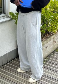 Ανοιχτό γκρί φόρμες με χαλαρή εφαρμογή, που διαθέτουν πλευρικές τσέπες και απαλή υφή. Φοριούνται με λευκά αθλητικά παπούτσια πάνω σε ξύλινη βεράντα.