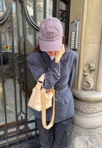 Personne portant une casquette violette, un blazer gris, un pantalon cargo foncé, portant un sac à bandoulière beige, debout près d'un pilier en pierre décoratif et d'un portail en fer forgé noir.