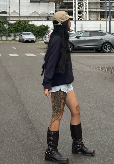 Botas negras hasta la rodilla con hebillas, pantalones cortos gris claro y una sudadera oscura. Cabello largo, tatuaje visible en la pierna. Entorno urbano.