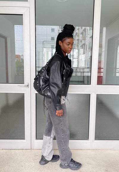 Chaqueta de cuero negra con grandes bolsillos, jeans de patchwork grises y zapatillas negras, peinada con trenzas y con un bolso negro. Ambiente interior.
