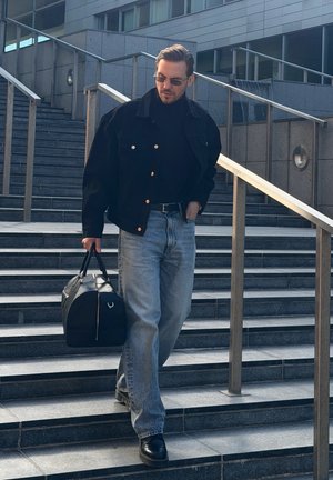 Hombre con gafas de sol, chaqueta negra y vaqueros bajando unas escaleras exteriores y llevando una bolsa de deporte negra en un entorno urbano.