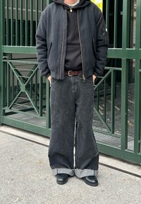 Blouson aviateur noir par-dessus un sweat à capuche gris, associé à un jean large noir avec des ourlets effilochés et des chaussures noires, debout près d'une porte verte.