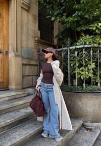 Brun tætsiddende top, cremefarvet lang frakke, lyseblå vide jeans, brun ruskinds håndtaske, grå sneakers, solbriller, stående ved en smedejernslåge.