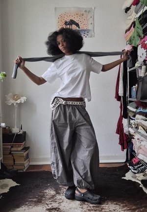 Mujer con pantalones cuadros sueltos, camiseta blanca y zuecos negros sosteniendo una bufanda gris estirada detrás de sus hombros en una habitación desordenada.