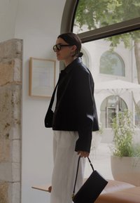 Kvinde i sort jakke og solbriller, der holder en sort håndtaske, står indendørs nær et vindue med planter og udendørs parasoller synlige.