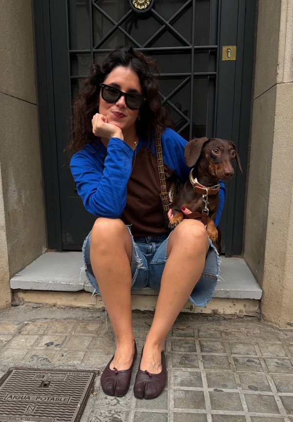 Mujer con gafas de sol y chaqueta azul sentada en un umbral sosteniendo un perro salchicha marrón, con pantalones cortos vaqueros rotos y zapatos con dedo dividido.